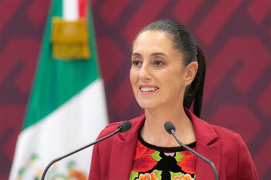 Claudia Sheinbaum giving a political speech with the Mexican flag in the background.