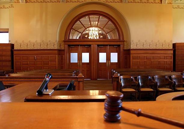 "An empty courtroom with wooden decor, featuring a judge's bench, a gavel, and a microphone, with chairs arranged for an audience and a chandelier hanging from the ceiling."